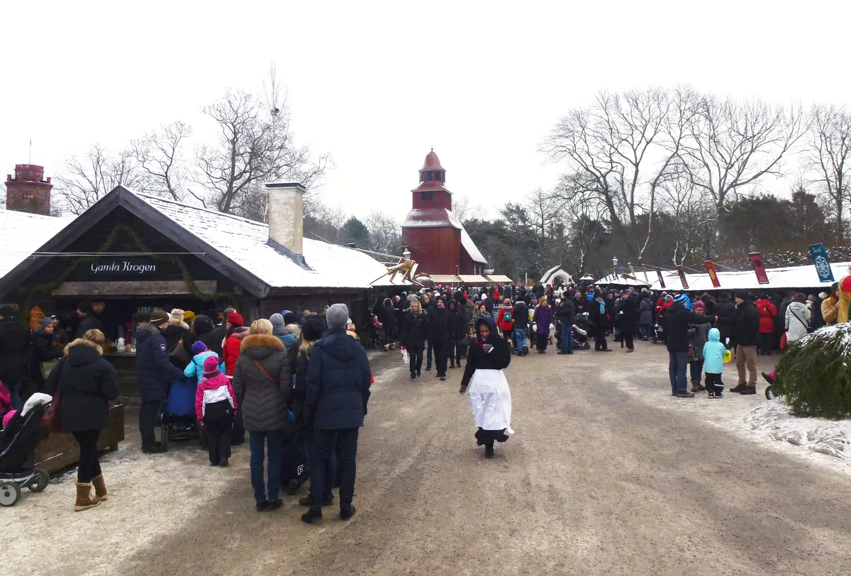 Besökare på Skansens julmarknad i snö bland de gamla trähusen